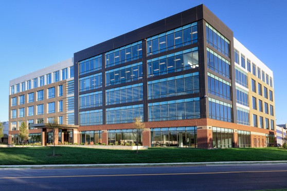 An office building with a brick first level, and black, brown and white panels on the other levels. Around the building is sidewalks and grass.