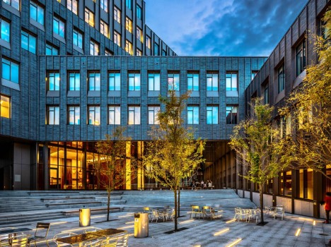 A modern building with a brick and glass facade. In front of the building is a patio with tiered seating, chairs and trees.