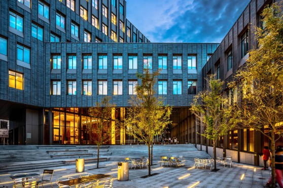 A modern building with a brick and glass facade. In front of the building is a patio with tiered seating, chairs and trees.