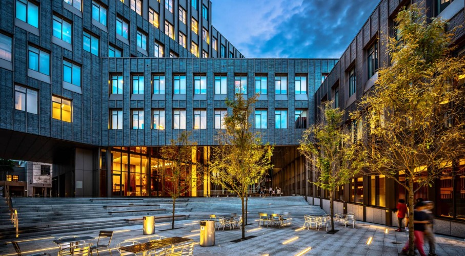 A modern building with a brick and glass facade. In front of the building is a patio with tiered seating, chairs and trees.