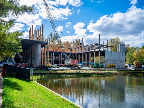 A crane sits in the middle of a concrete building with wooden colums. A lake is in front of the site, and many other machines and building materials are seen around the job.