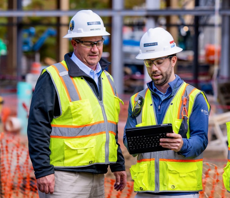 3 men in yellow safety vests and hard hats look at an iPad on a job site.