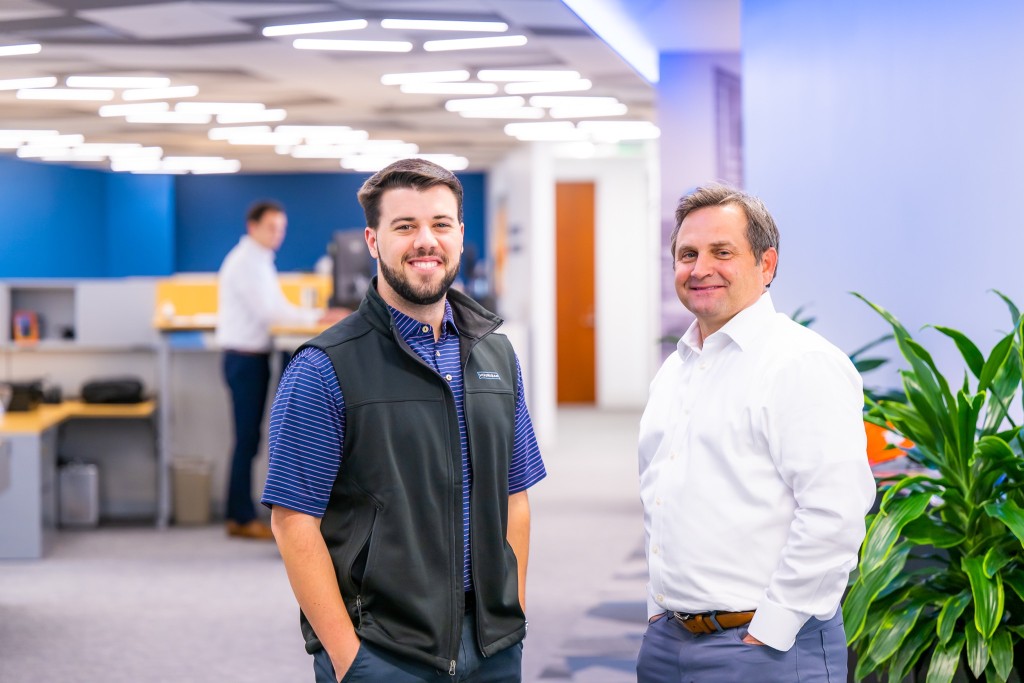 Two men in business attire pose in front of an office.