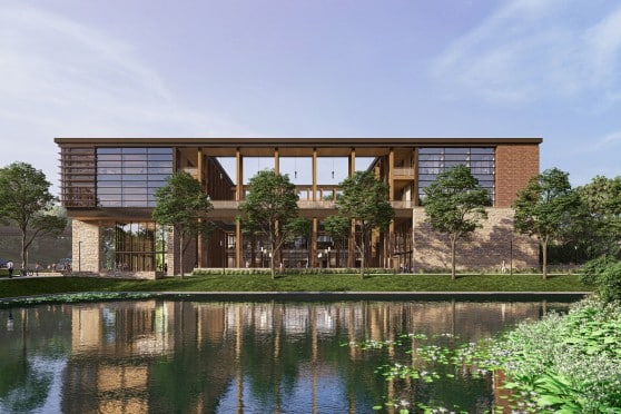 Image depicts a modern black metal building with wooden beams and windows. There is a body of water and plants in the foreground.