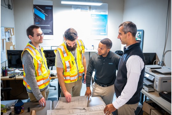 4 people, two of which are wearing safety vests, look over construction documents in a small room.