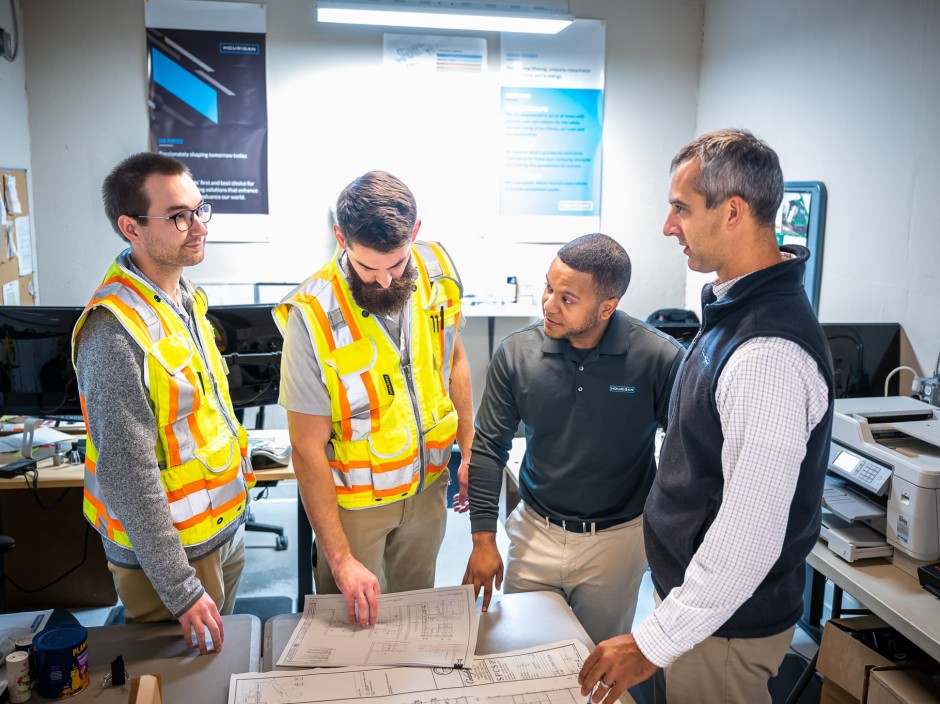4 people, two of which are wearing safety vests, look over construction documents in a small room.