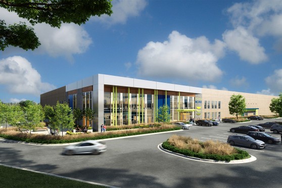 Modern two-story factory building with glass facade and blue and green accents under a clear blue sky. Cars are in the parking lot that sits in front of the building.