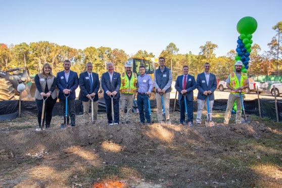 10 people hold shovels in the ground and smile for the camera at a groundbreaking. Behind them is a construction site.