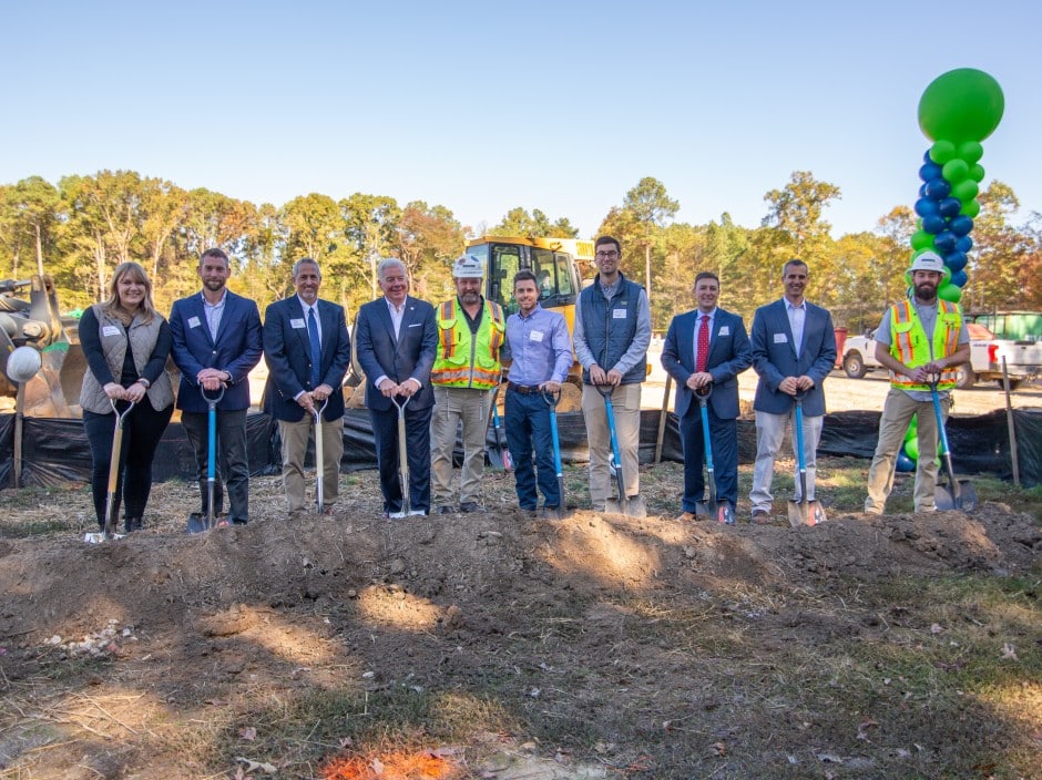 10 people hold shovels in the ground and smile for the camera at a groundbreaking. Behind them is a construction site.