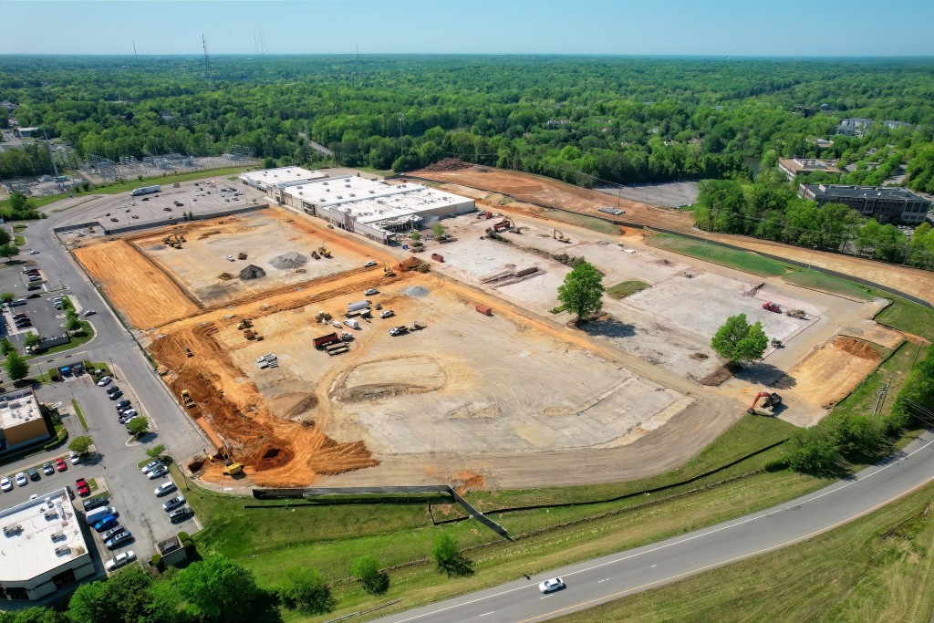 An aerial view of a dirt building pad surrounded by trees, roads and other buildings.
