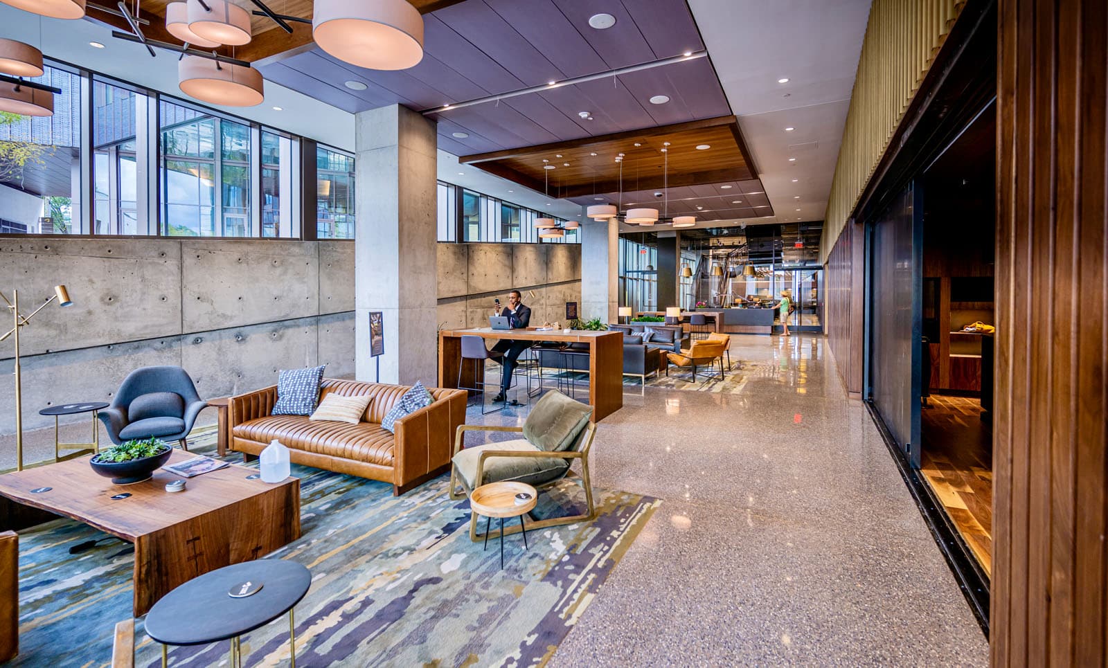 Modern lobby area with a mix of concrete walls, wooden paneling, and glass windows. The space includes a seating area with sofas and chairs, a desk with a person working on a laptop, and a reception desk.