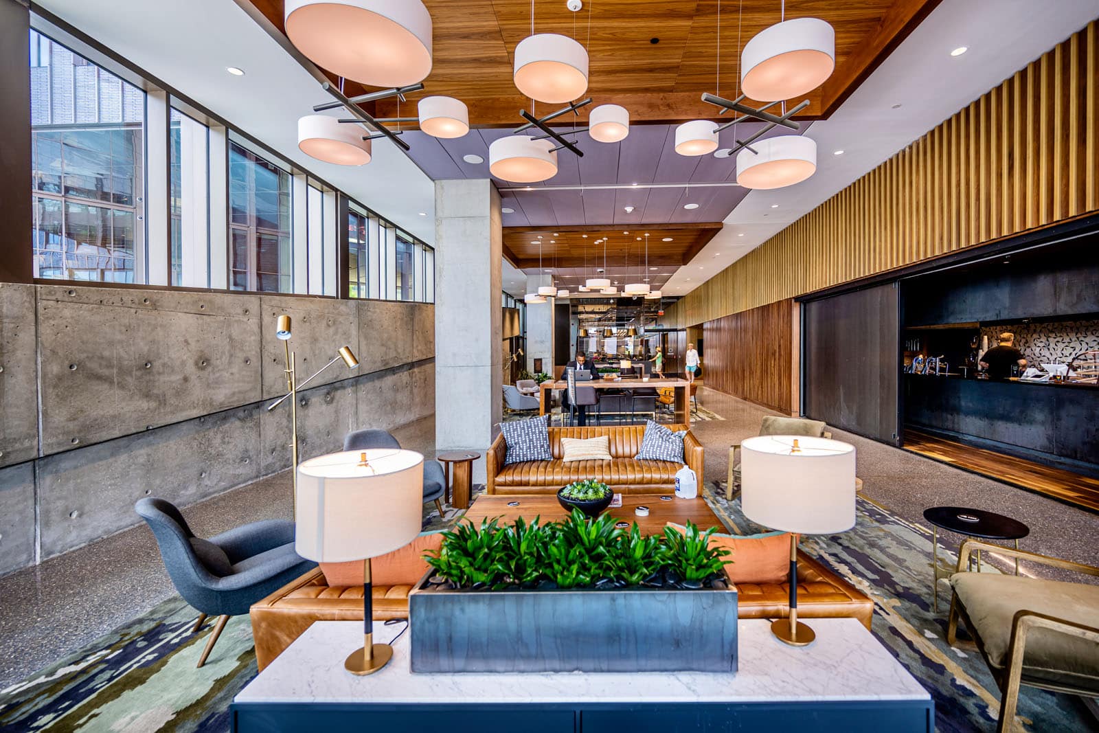 A modern lobby with a mix of contemporary furniture including sofas, armchairs, and tables under warm lighting. The space features wooden ceiling panels, concrete walls, and a view to an adjacent area through a glass partition.