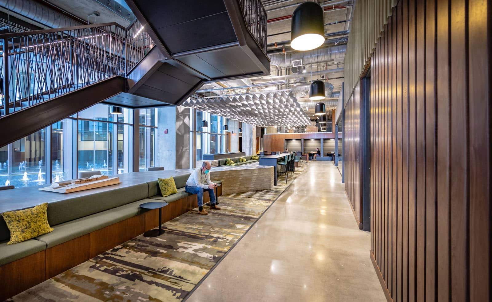 A man sit on a couch in a lobby with a blend of eclectic furniture. Tan sofas, yellow armchairs, and a dining area are visible. The ceiling features an intricate geometric pattern, and large windows allow ample natural light.