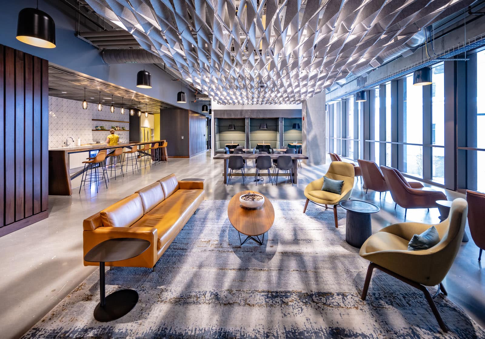 The image depicts a modern and spacious interior with a blend of eclectic furniture. Tan sofas, yellow armchairs, and a dining area are visible. The ceiling features an intricate geometric pattern, and large windows allow ample natural light.