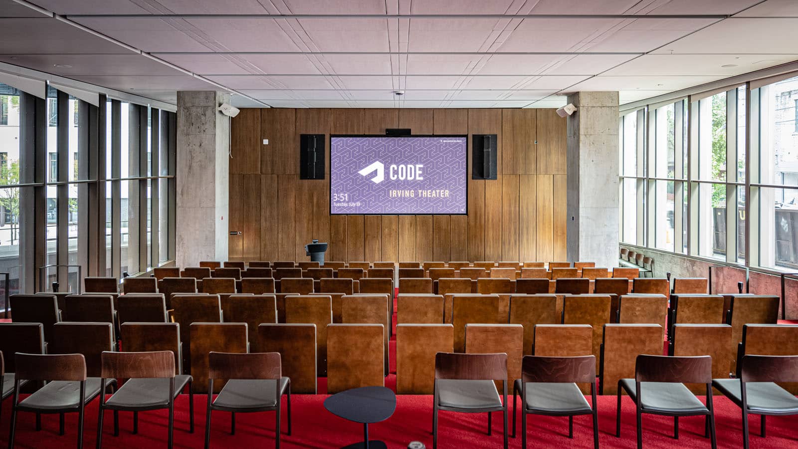 An auditorium with many wood chairs facing a wood paneled wall and screen. The floors have red carpeting, and the walls are concrete and wood.