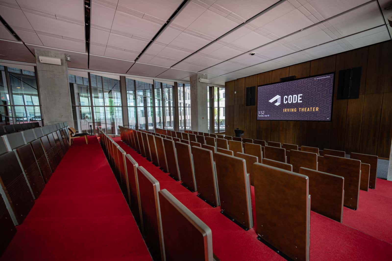 An auditorium with many wood chairs facing a wood paneled wall and screen. The floors have red carpeting, and the walls are concrete and wood