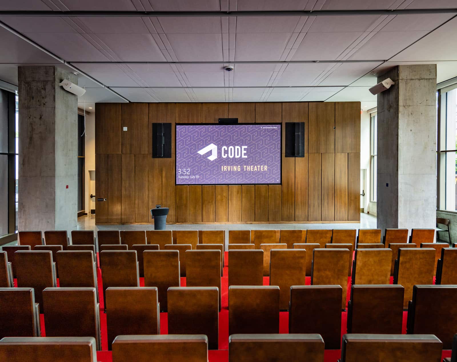 An auditorium with many wood chairs facing a wood paneled wall and screen. The floors have red carpeting, and the walls are concrete and wood