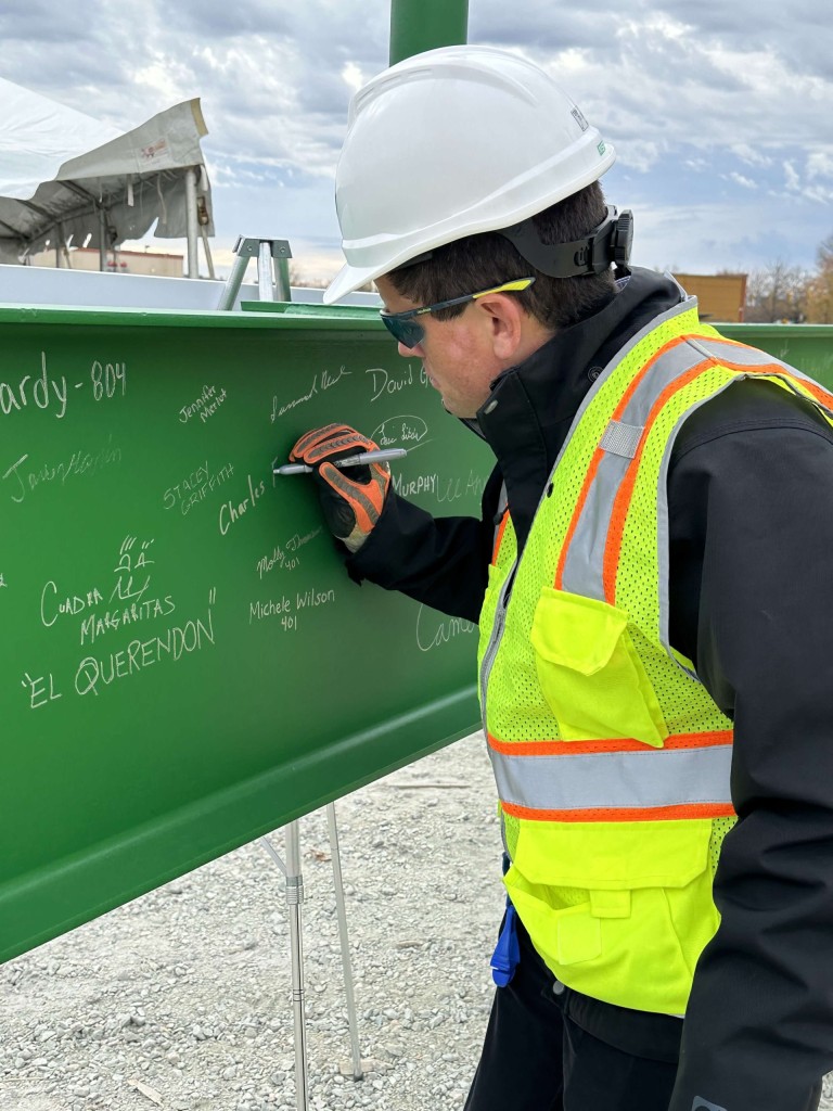 Man in black shirt, yellow construction vest and white hardhat sighs a large green metal beam.
