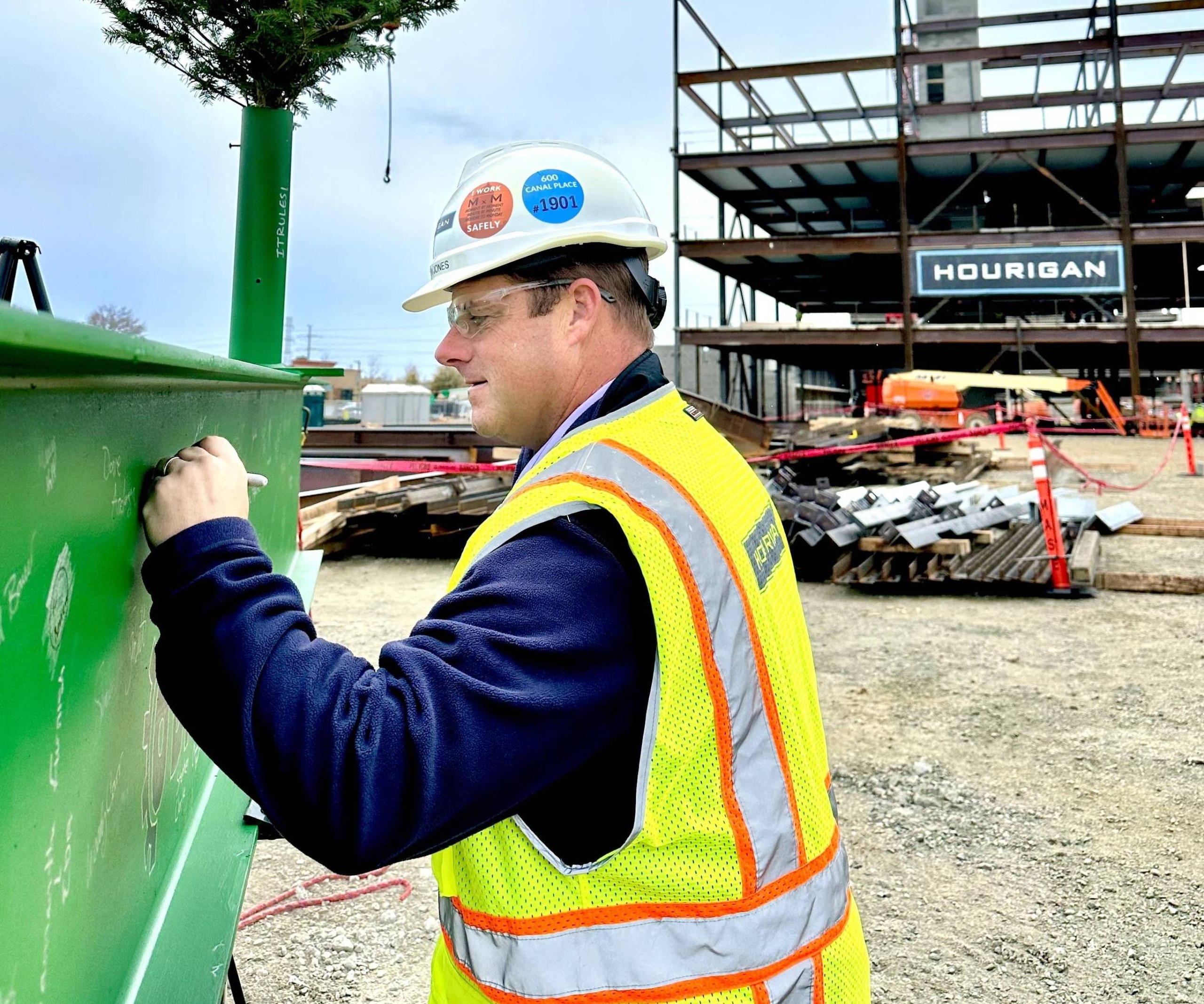 A man in a blue sweater wears a hard hat and yellow safety vest to sign a large green construction beam in front of a building under construction.