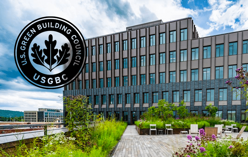 A modern multi-story building with a geometric facade, surrounded by garden with wooden pathways and purple flowers under a cloudy sky.
