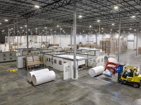 Image depicts a factory with shelves full of paper rolls in the background. Teh foreground depicts paper processing machinery and a yellow forklift picking up a large roll of whiter paper.