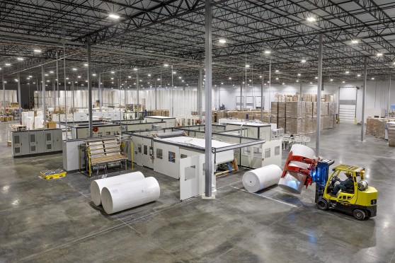 Image depicts a factory with shelves full of paper rolls in the background. Teh foreground depicts paper processing machinery and a yellow forklift picking up a large roll of whiter paper.