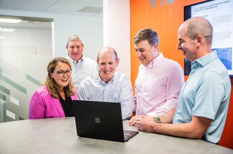 5 people stand around a laptop and laugh